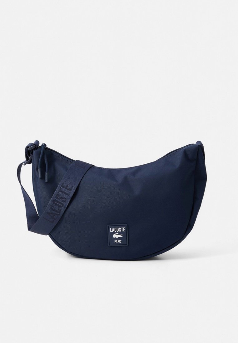 Sac bandoulière bleu marine en forme de croissant avec une sangle réglable embossée "Lacoste" et un logo patch à l'avant indiquant "Lacoste Paris" avec un crocodile.
