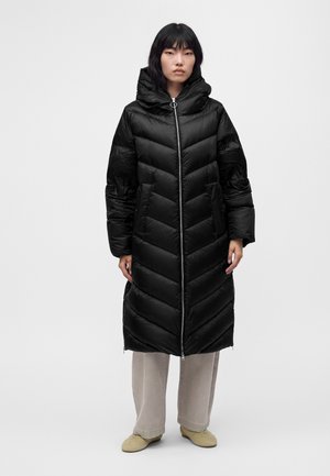 Femme debout portant un long manteau matelassé noir avec capuche, un pantalon beige à jambes larges et des chaussures beige clair sur fond blanc.