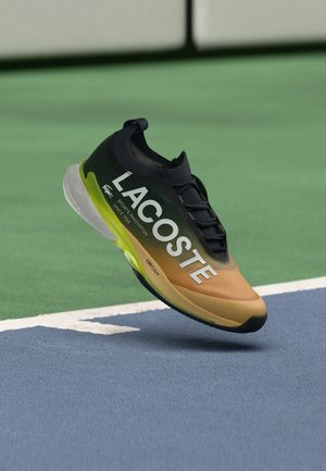 Scarpa sportiva Lacoste nera e marrone con testo bianco e accenti verdi, che fluttua sopra una superficie di campo da tennis blu e verde.