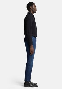 Camicia a maniche lunghe scura abbinata a jeans blu, che mette in evidenza una vestibilità slim e la trama del denim. Stivali neri alla caviglia completano l'outfit.