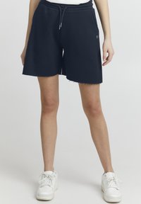 Marineblå bomuldsbukser med elastisk talje og snørre, med en frynset kant og sidelommer. Bæret med hvide sneakers.