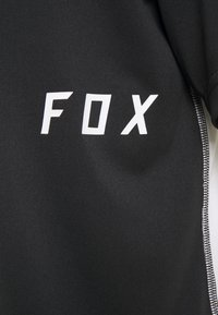 Černé sportovní tričko s bílým nápisem "FOX". Texturovaná látka s aerodynamickým designem a kontrastním bílým prošitím podél švů.