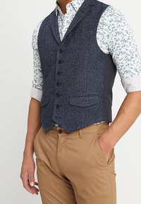 Donkerblauwe textuurvest met vier knopen, twee voorzakken en grijze zijpanelen, gedragen over een bloemenpatroon shirt en tan broek.