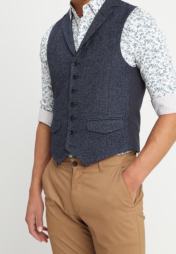 Donkerblauwe textuurvest met vier knopen, twee voorzakken en grijze zijpanelen, gedragen over een bloemenpatroon shirt en tan broek.