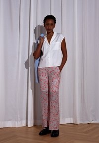 Blusa branca sem mangas com um bolso, combinada com calças largas de padrão floral vermelho. Sapatos pretos. O fundo apresenta cortinas brancas.