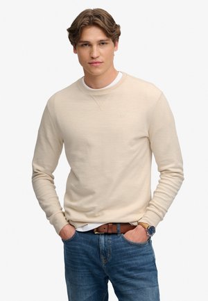 Ung mand iført en beige sweater, blå jeans, brun bælte og armbåndsur, stående med hænder i lommerne mod en hvid baggrund.