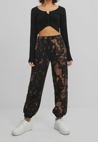 Svart långärmad crop top med knappr detaljer, som bärs tillsammans med löst sittande svarta och bruna tie-dye joggers med elastiska muddar och dragsko i midjan.