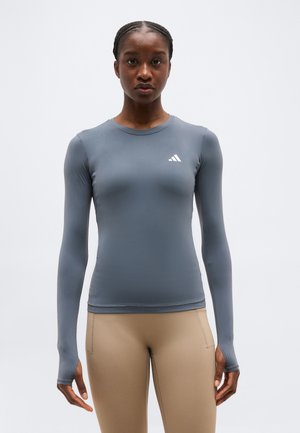 Žena oblečená v priliehavom sivom športovom tričku s dlhým rukávom a otvormi na palce a béžových legínach, stojaca na jednoduchom svetlom pozadí.