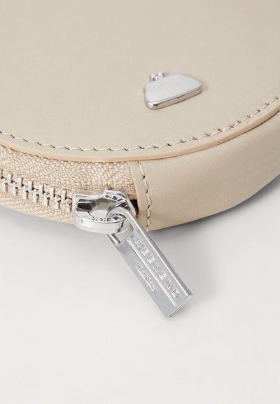 Bolsita redonda de cuero beige con herrajes plateados y un tirador de cremallera texturizado. Presenta un pequeño emblema en la superficie.