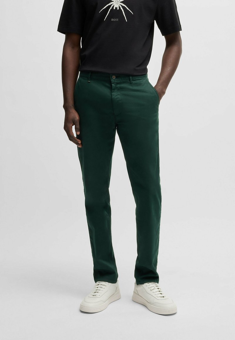 Ciemnozielone spodnie chino z gładkiego materiału, o zwężanym kroju, z standardowymi kieszeniami, w zestawieniu z białymi trampkami i czarnym t-shirtem.