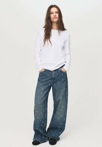 White long-sleeve shirt paired with loose-fitting blue denim jeans featuring a white circular pattern. Model stands with hands in pockets.