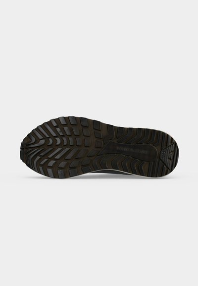 Semelle de chaussure en caoutchouc noir avec des motifs de bande de roulement profonds et variés et une légère courbe, présentée sur un fond gris clair uni.