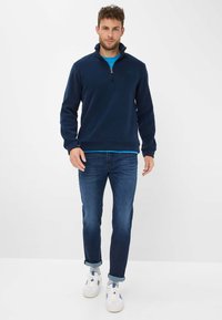 Pullover blu scuro con colletto a mezza zip, morbida texture in pile, indossato sopra una camicia azzurra chiara, abbinato a jeans scuri e sneakers bianche.