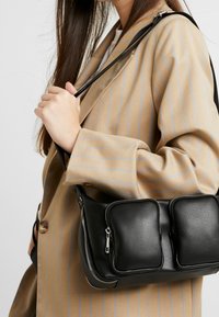 Borsa a mano in pelle nera con due tasche frontali con cerniera, texture liscia e una tracolla robusta e regolabile, tenuta da una persona con un cappotto beige a righe.