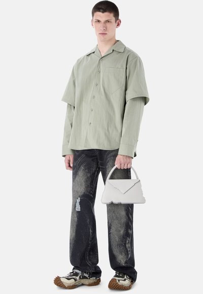 Jeune homme portant une chemise à boutons vert sauge superposée et un jean noir déchiré, tenant un petit sac à main blanc, chaussé de baskets épaisses.
