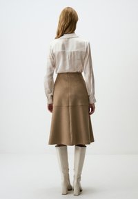Jupe mi-longue en daim beige associée à un chemisier à motifs crème et marron clair et des bottes blanches à talons blocs montantes.