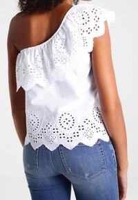 White cotton top with one shoulder design, featuring a scalloped eyelet lace trim and embroidered floral patterns along the hem.