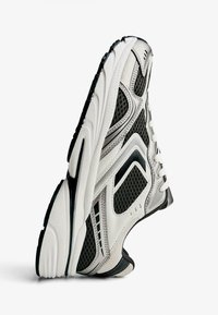 Hvid og sort sportsko med mesh-paneler, struktureret sål, polstret krave og kontrasterende detaljer på en minimal baggrund.
