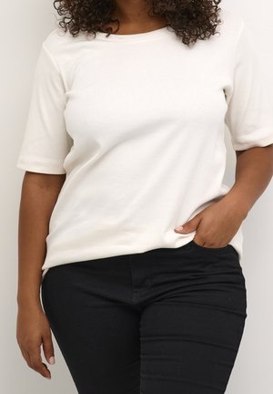 Person with curly hair wears a white short-sleeve top and black pants, with one hand in pocket and standing against a plain background.