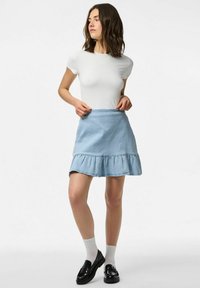 Šviesiai mėlynos džinsinės sijonas su raukšlėtu kraštu, derinamas su baltu prigludusiu marškinėliu ir juodais loaferiais, dėvimi virš baltų kulkšninių kojinių.