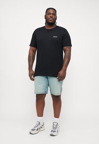 Schwarzes, lässiges T-Shirt aus Baumwolle mit kurzen Ärmeln, ausgestattet mit einem kleinen weißen Logo. Kombiniert mit hellblauen Denim-Shorts und Sneakers.
