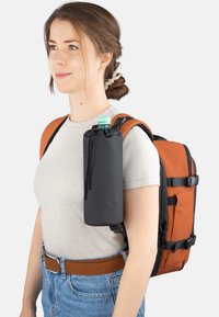 Oranger Rucksack mit schwarzen Gurten; befestigter schwarzer Flaschenhalter; glatter Stoff; zylindrische Flasche mit grünem Deckel; legere Kleidung.