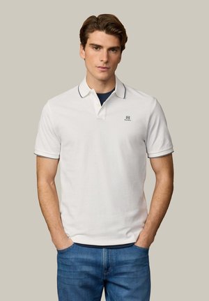 Jonge man met een wit poloshirt met korte mouwen, navy rand en logo, gecombineerd met een blauwe spijkerbroek, staand met handen in zijn zakken.