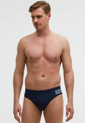 Hemdloser Mann mit kurzen hellbraunen Haaren, der dunkelblaue Badehosen mit weißem "Camp David"-Schriftzug an der linken Hüfte trägt.