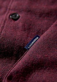 Bordeaux gekleurde textuurstof met een knop en een blauwe label met de tekst "LINDBERGH." De stof heeft een subtiel patroon en een gladde afwerking.