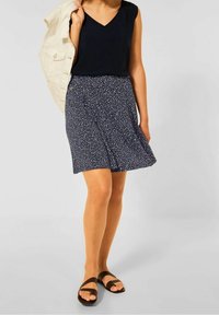 Tmavě modrá sukně s bílým puntíkovým vzorem, spárovaná s černým tílkem bez rukávů. Modelka má na nohou světle hnědé sandály a drží světlou bundu.