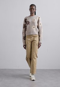 Strickpullover in hellem Beige mit braunen und cremefarbenen Blumenmustern, kombiniert mit khakifarbenen Cargo-Hosen. Model trägt silberne Sneaker.