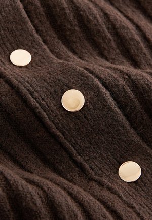 Brūns teksturēts audums ar vertikālām ribām un trim izciliem zelta toņa pogām. Materiāls šķiet mīksts un silts.