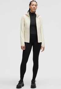 Light cream zip-up jacket with a hood over a black long-sleeve top and black leggings, featuring a fitted design and textured fabric.