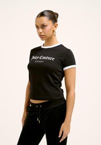 Černé krátké tričko s bílým lemem, na kterém je v tučném písmu nápis "Juicy Couture SPORT". Model má na sobě černé sametové tepláky se zlatými akcenty.