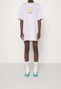 Lila oversized t-shirt med en färgglad grafisk bild av en sol och moln, i kombination med teal sneakers och randiga strumpor. En enfärgad bakgrund.