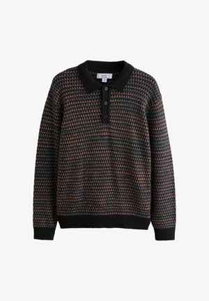 Mann steht mit den Händen in den Taschen, trägt ein gemustertes langärmeliges Polo, graue plissierte Hose und braune Lederschuhe vor beigem Hintergrund.