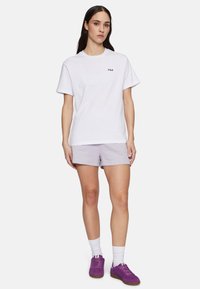 T-shirt bianca in cotone con logo Fila, abbinata a pantaloni corti lavanda, calze bianche a crew e sneakers in suede viola. Insieme semplice e informale.
