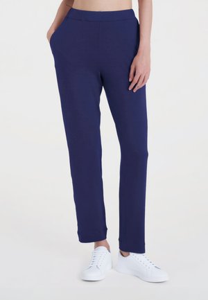 Donna con pantaloni blu navy a gamba dritta e sneakers bianche, in piedi con una mano in tasca contro uno sfondo semplice.