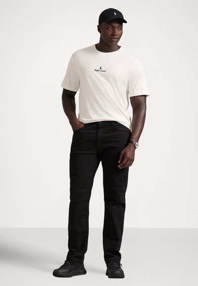 Hvit t-skjorte med korte ermer og svart logo, svarte jeans og svarte joggesko. Modellen har på seg en svart caps og står med hendene i lommene.