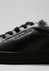 Baskets en cuir noir avec lacets noirs, logo Calvin Klein embossé sur le côté, et doublure en fourrure grise visible sur le bord supérieur.
