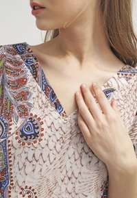 Top de cuello en V con intrincados patrones de paisley multicolores sobre una tela ligera con un fondo marrón texturizado, que presenta un diseño de media manga.