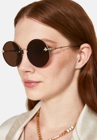 Gafas de sol redondas marrones con montura de metal dorado y acento de abeja en la patilla. Lente suave y diseño elegante. Escote texturizado visible.