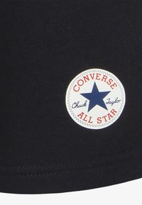 T-shirt en tissu noir arborant un logo circulaire avec une étoile bleue, du texte rouge indiquant "CONVERSE ALL STAR" et "Chuck Taylor" en police cursive.