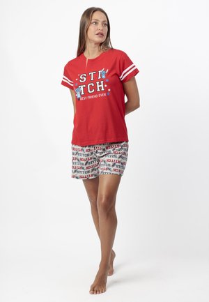 Mujer con camiseta gráfica roja de Stitch y pantalones cortos blancos con estampado de texto Stitch, de pie descalza sobre fondo blanco.
