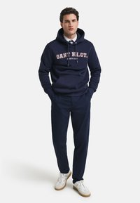 Dunkelblaue Kapuzenjacke mit weißem und rotem Text, mit einer vorderen Taschentasche, kombiniert mit dunkelblauen Hosen und weißen Sneakern. Glattes Material, lässige Passform.