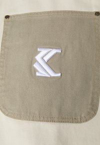 Beige stofflomme med en tykk sømkant, som har en hvit brodert logo som ligner på en sammenflettet "K"-form.