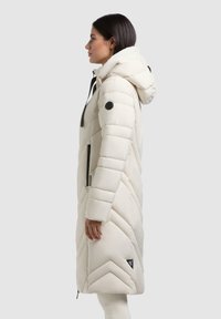 Giacca piumino leggera di colore bianco sporco, dotata di cappuccio, trapuntatura a zig-zag, tasche con zip e una toppa nera con logo sulla manica.