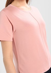 Jasnoróżowa koszulka z krótkim rękawem, z okrągłym dekoltem, gładką fakturą i subtelnymi detalami po bokach. Wykonana z miękkiego, elastycznego materiału.