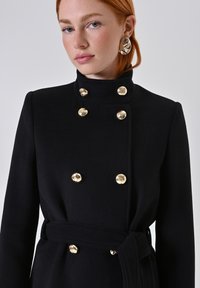Zwarte dubbelgetailleerde jas met een staande kraag, gouden knopen en een bijpassende riem. Heeft een getailleerde pasvorm en een zachte stoftextuur.