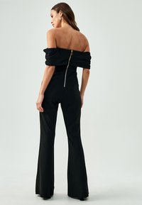 Sorte off-the-shoulder jumpsuit med tætsiddende overdel, lange flagrende ben og baglynlås. Blød tekstur, minimal detaljer.
